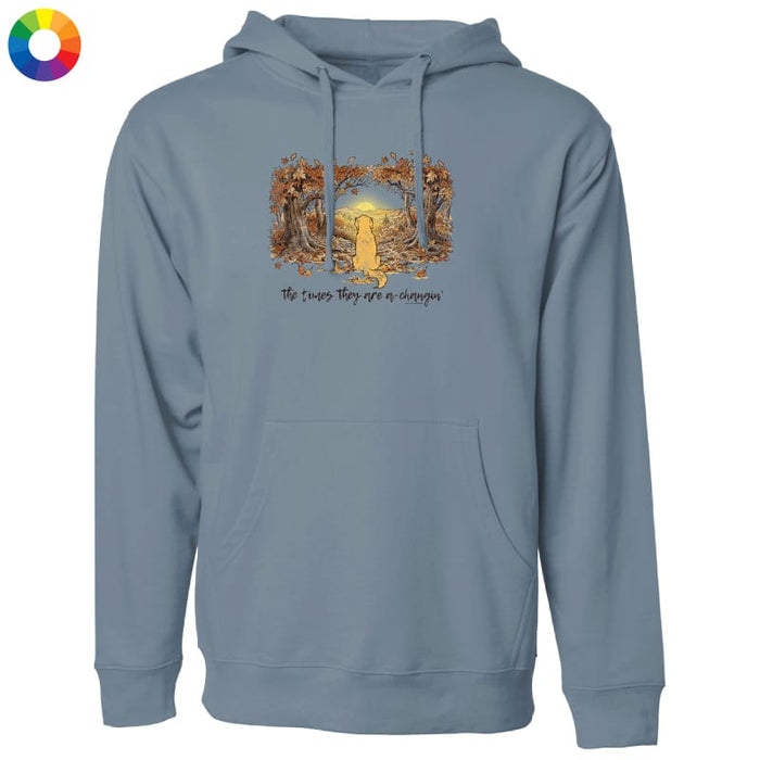 A dusty blue-gray pullover hoodie with a front kangaroo pocket and drawstring hood, featuring a central graphic of a yellow dog sitting in a field framed by autumn trees, with the text 'The dogs they love are everyone'. A small multicolor color wheel icon is in the top-left corner on a white background.