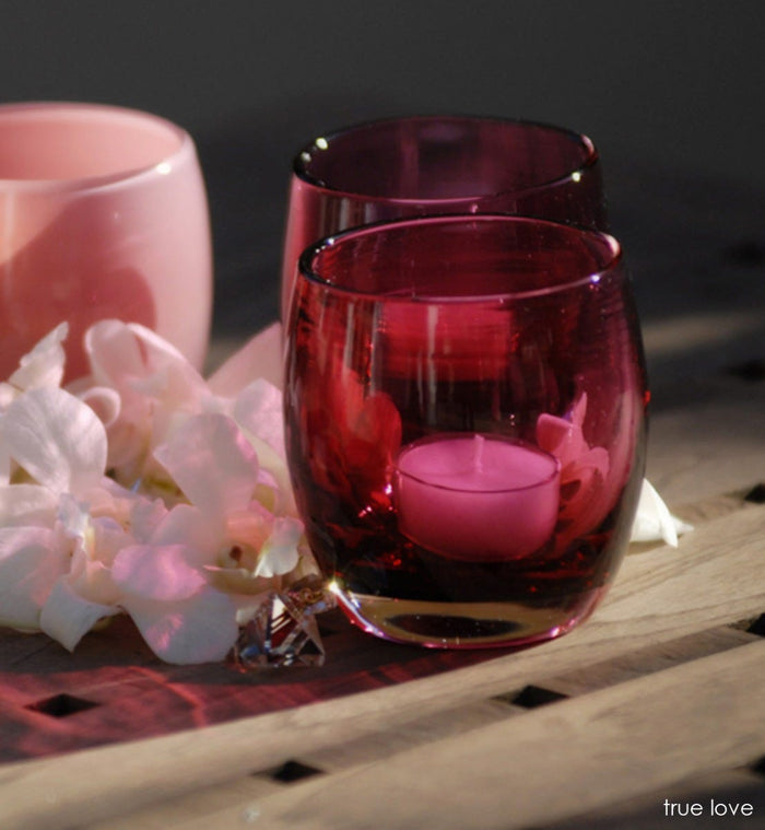 two true love, transparent deep raspberry pink, hand-blown glass votive candle holders next to white flowers.