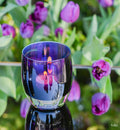 tulip, dark purple with silver metallic interior, hand-blown glass votive candle holder on an outdoor reflective surface with purple tulips in the background.