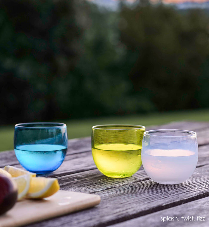 twist drinker, transparent lime green, hand-blown drinking glass with splash and fizz all filled with liquid on wood picnic table next to a cutting board with lemons.