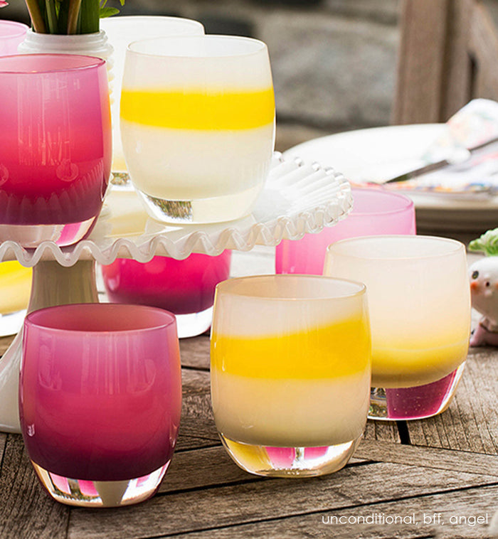 unconditional, white with yellow stripe, hand-blown glass votive candle holder. Paired with bff and angel on an outdoor wood table with flowers and a cake tray.