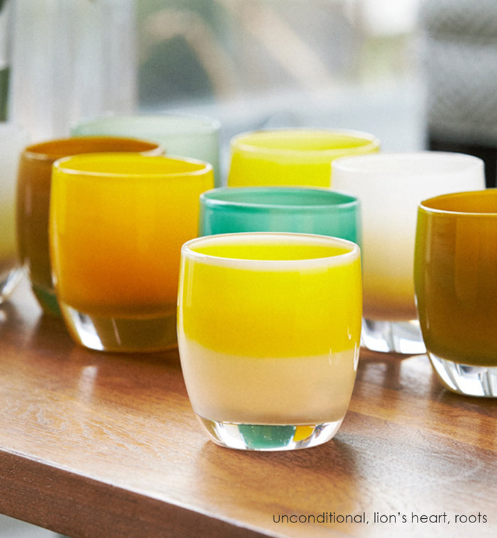 unconditional, white with yellow stripe, hand-blown glass votive candle holder on a light wood table with grouping of lion's heart and roots and peaceful.