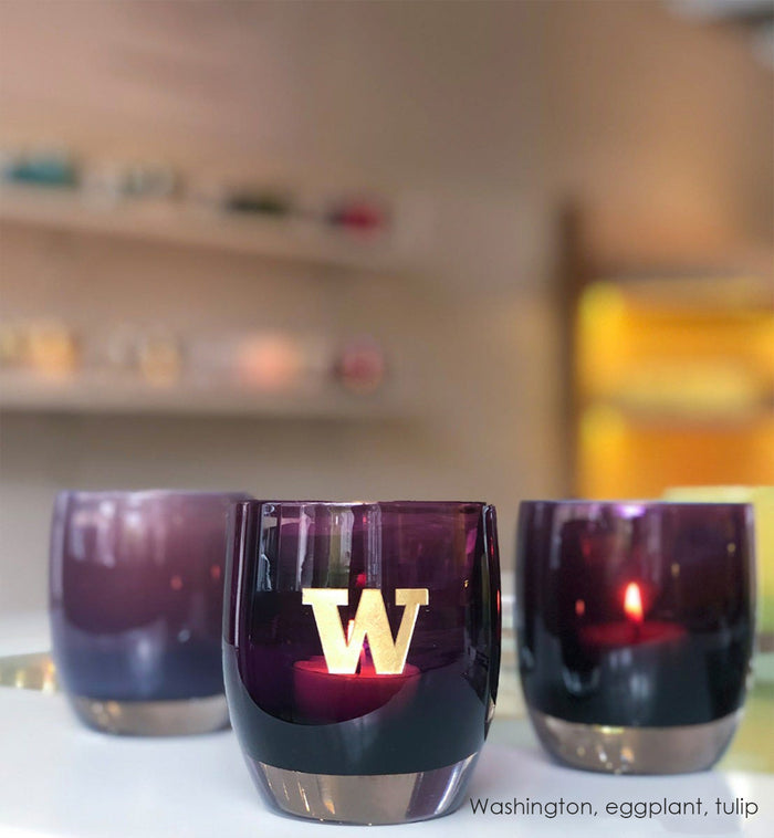 Washington, purple with sandblasted university of washington etching hand painted in gold, hand-blown glass votive candle holder. Paired with eggplant and tulip on a white table in a glassybaby retail store.