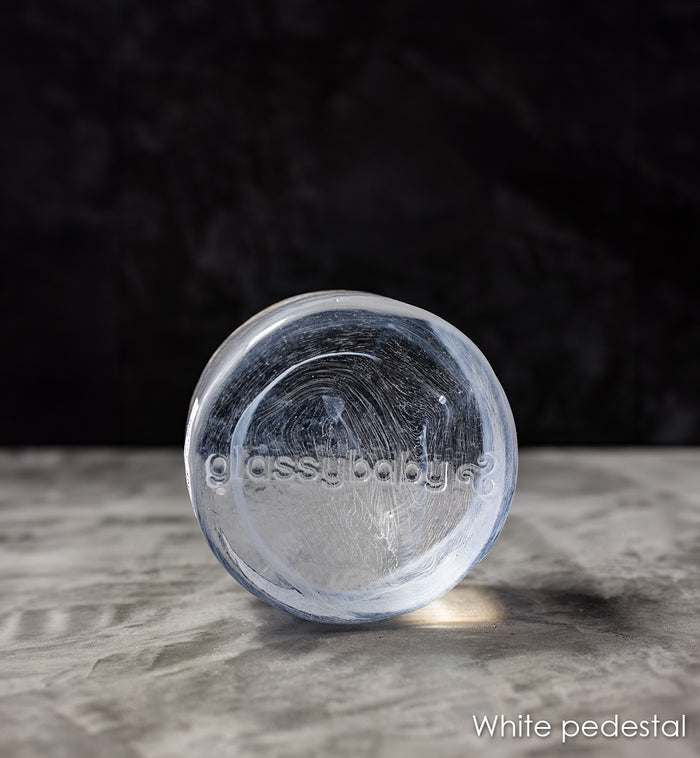 white transparent glass pedestal with 'glassybaby' branding on a gray surface with a dark background
